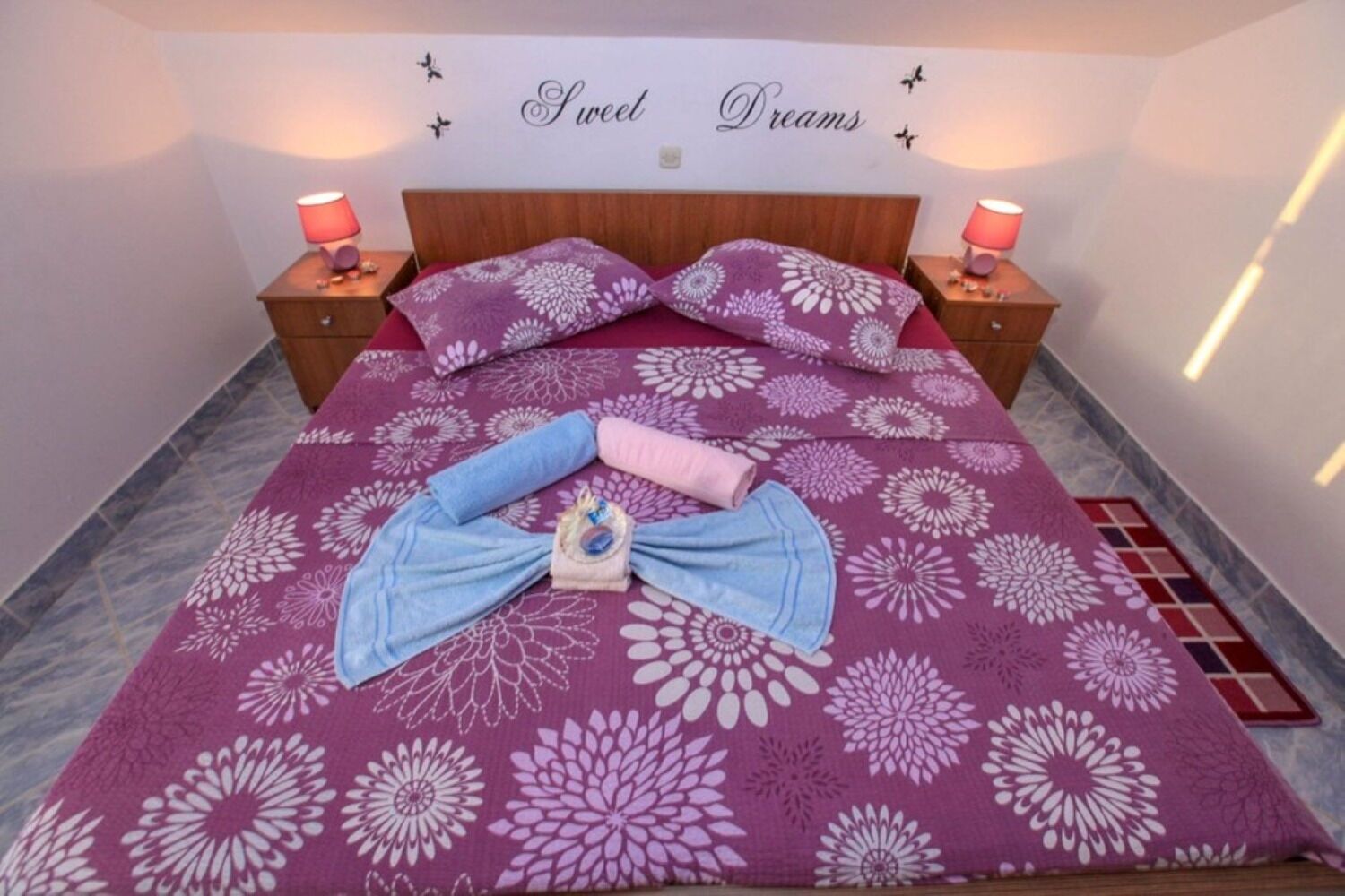 A neatly made bed with a floral bedspread, two pillows, and a towel arrangement on a tiled floor.
