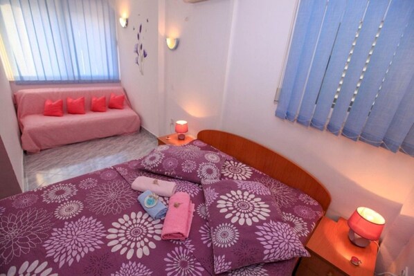 A bedroom with a bed, a bedside table, a lamp, a window with blinds, and a sofa with cushions. - Apartments Drago (Slivno)