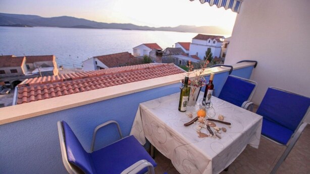 A balcony with a table set for two, overlooking a body of water and buildings. - Apartments Drago (Slivno)