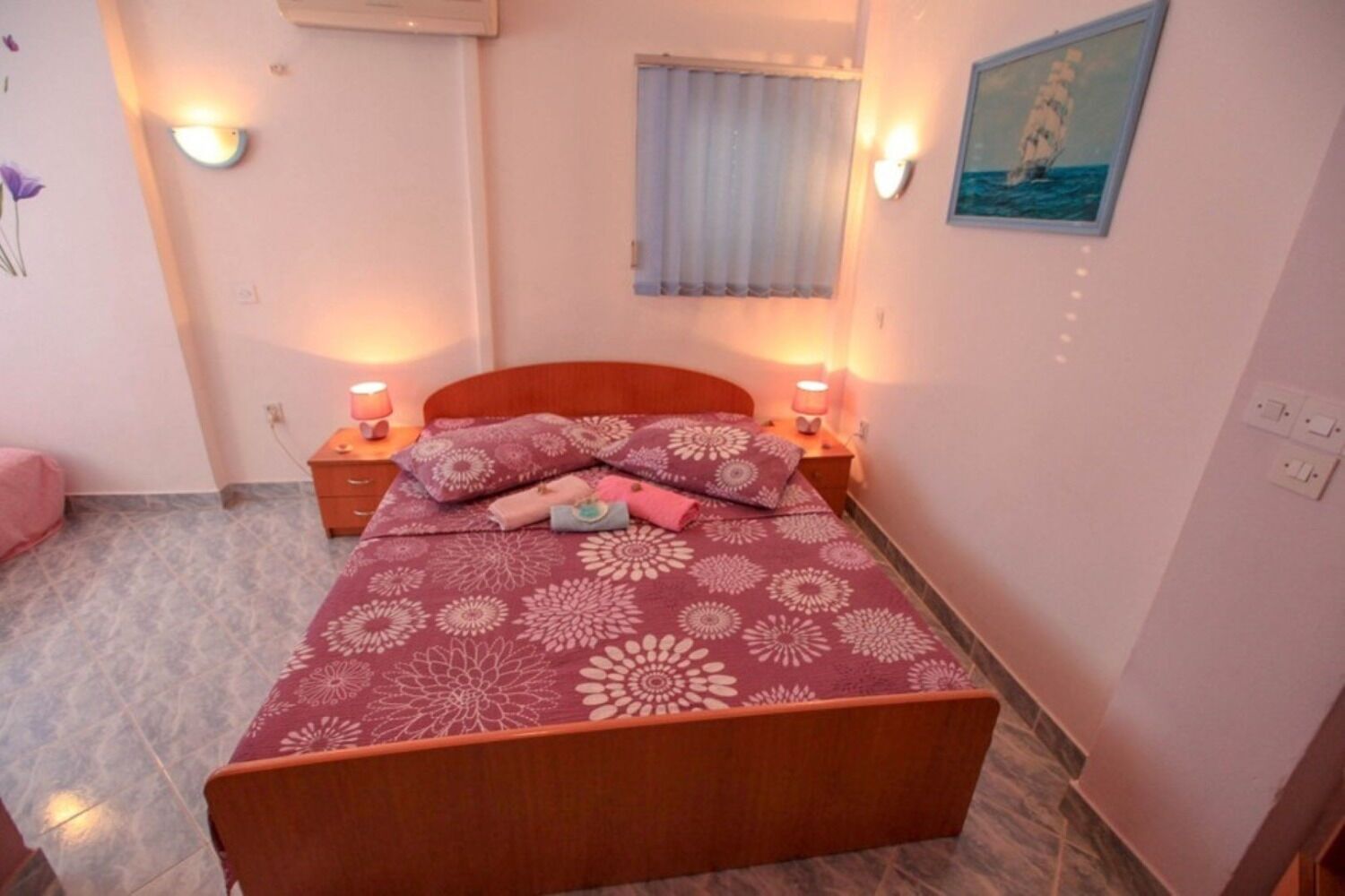 A bedroom with a bed, two bedside tables, a window with curtains, and a wall-mounted air conditioner.