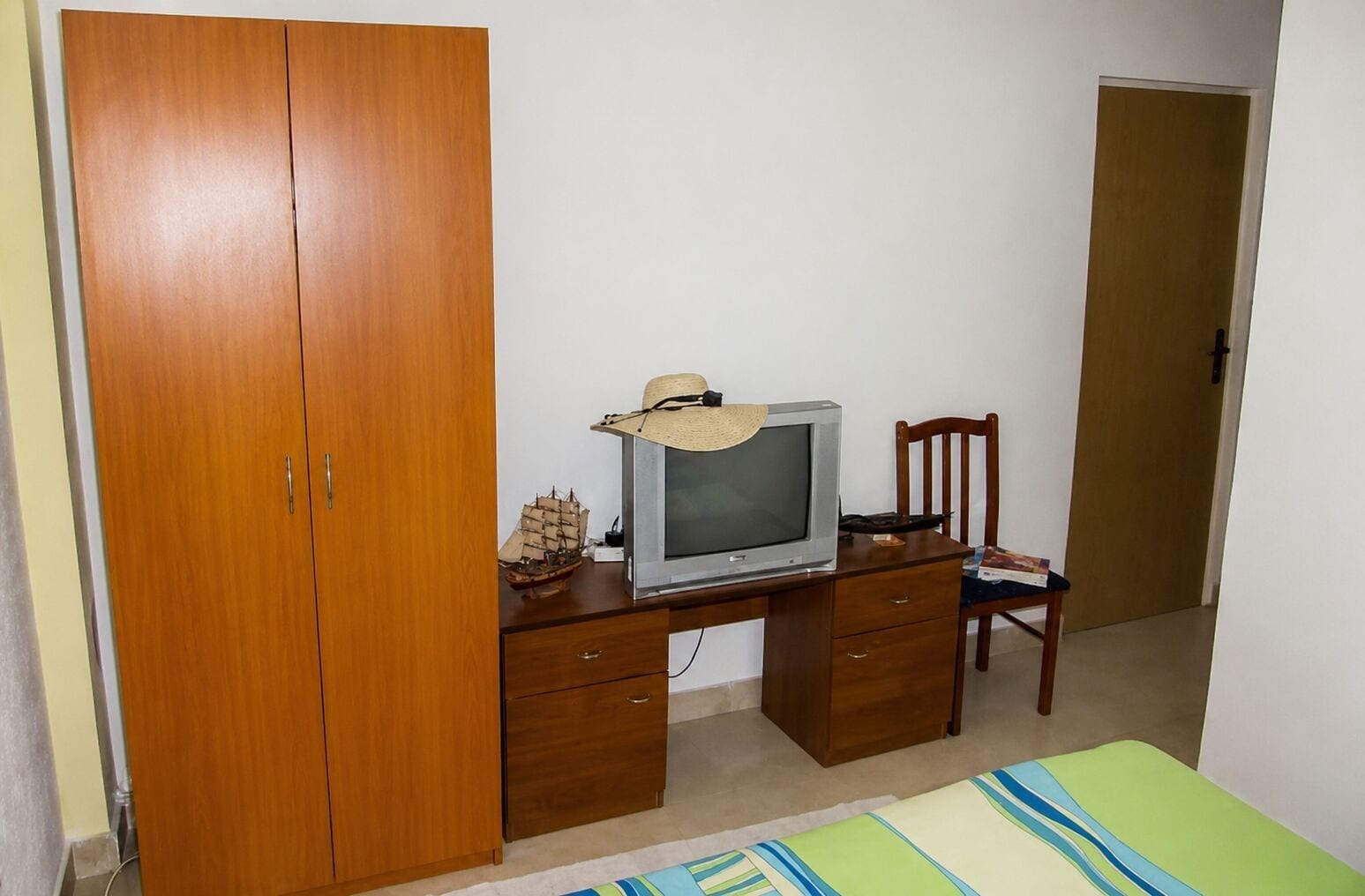 A bedroom with a wooden wardrobe, a television on a wooden desk, a small ship model, a straw hat, and a green-striped bedspread.