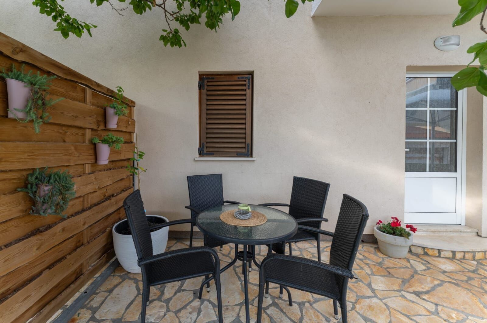 A patio with a round table and four chairs, a wooden wall with potted plants, and a window with blinds.