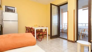 Ein Zimmer mit einem Bett, einem Esstisch mit Stühlen, einem kleinen Tisch mit einer Schüssel, einem Kühlschrank und einer Glastür, die auf einen Balkon mit Sitzgelegenheiten führt.