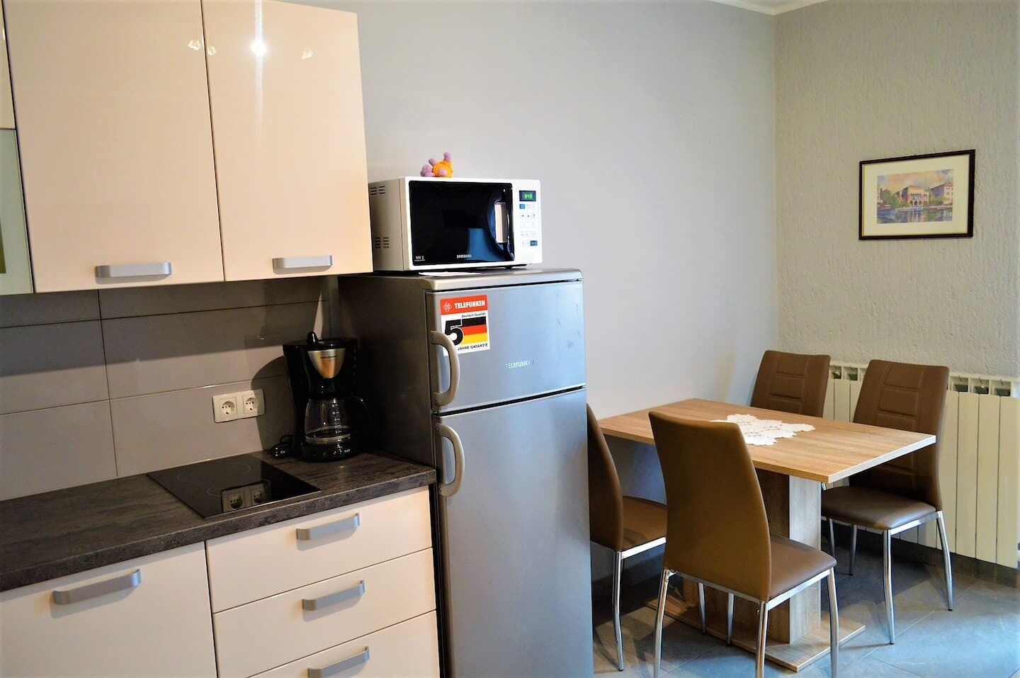 Een compacte keuken met een magnetron, koffiezetapparaat en een koelkast. Er is een eettafel met stoelen en een ingelijste foto aan de muur.