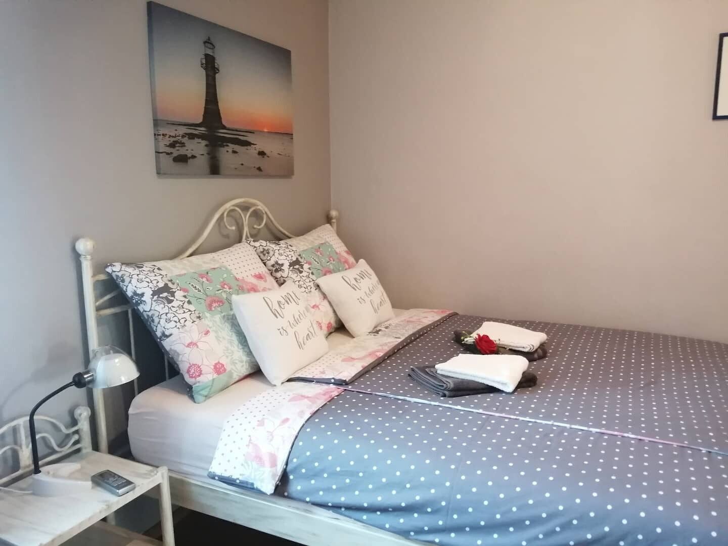 A neatly made bed with a polka dot bedspread, floral pillows, and a small bedside table with a lamp and a phone.