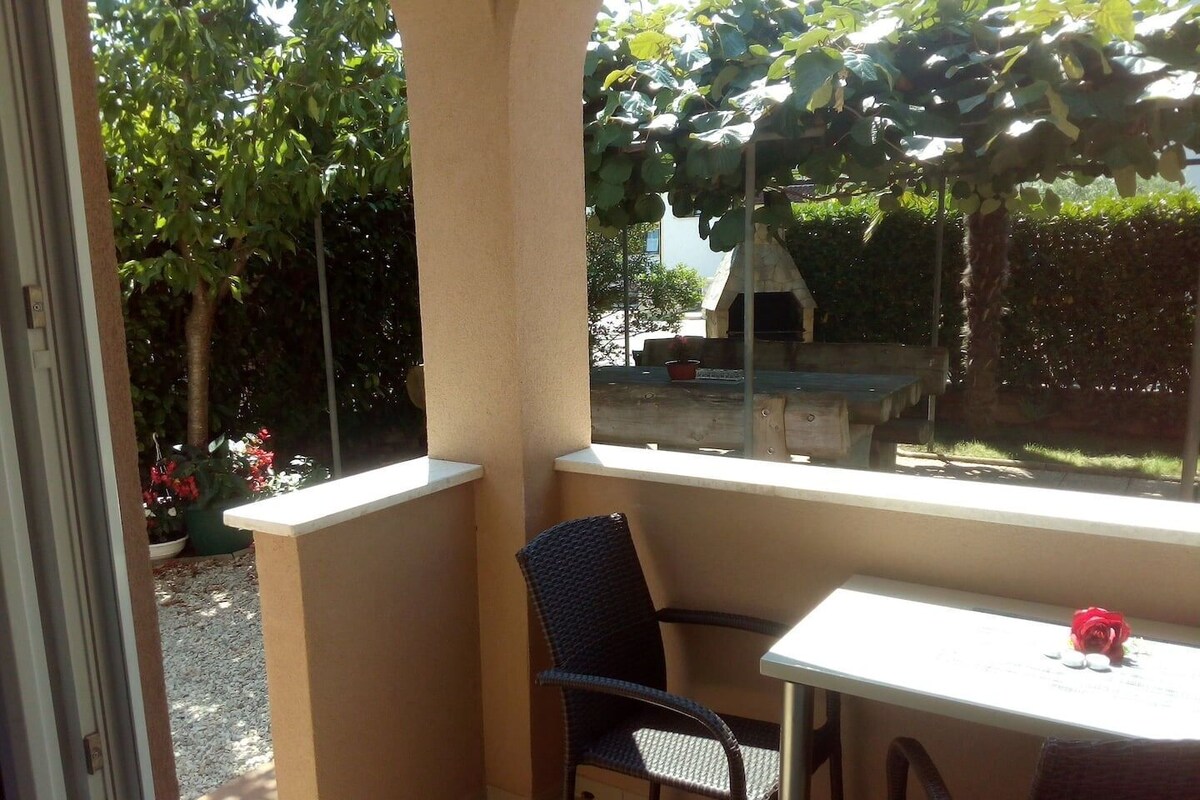 A balcony with a table and two chairs, overlooking a garden with a barbecue area and a tree.