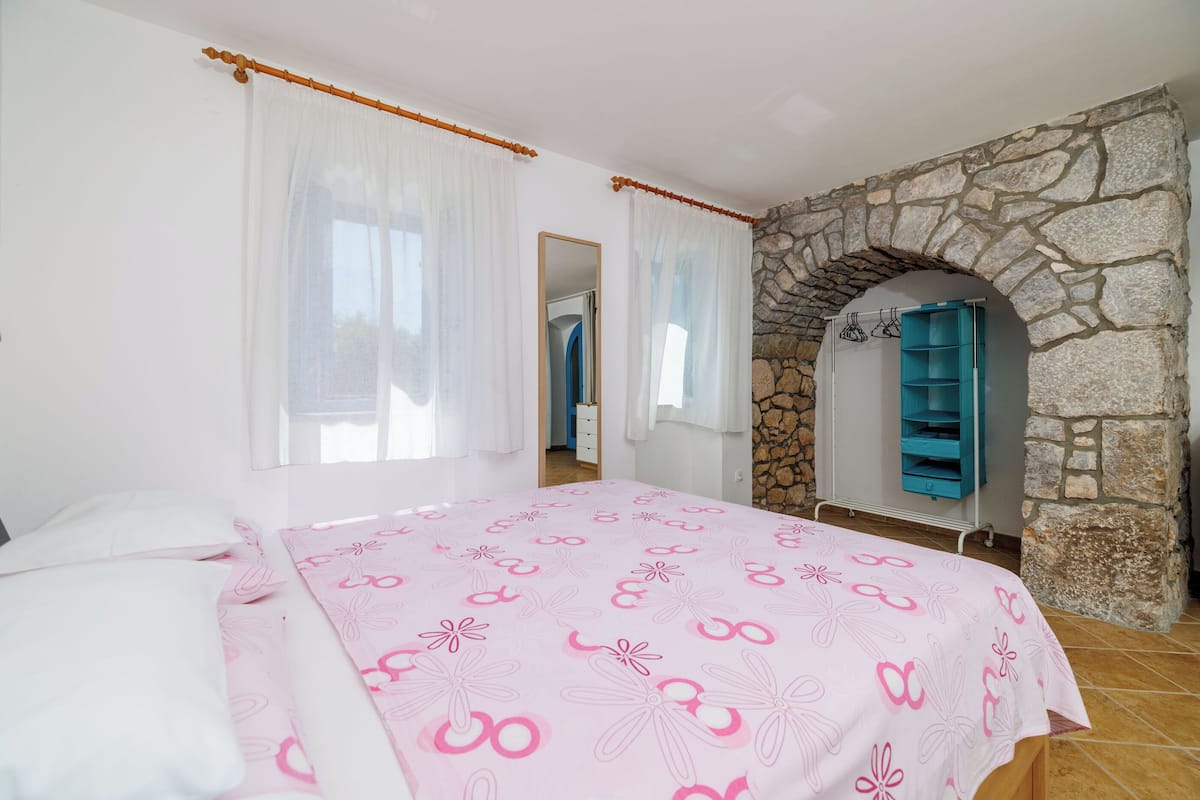 A bedroom with a bed, white curtains, a stone wall, and a blue shelf unit.
