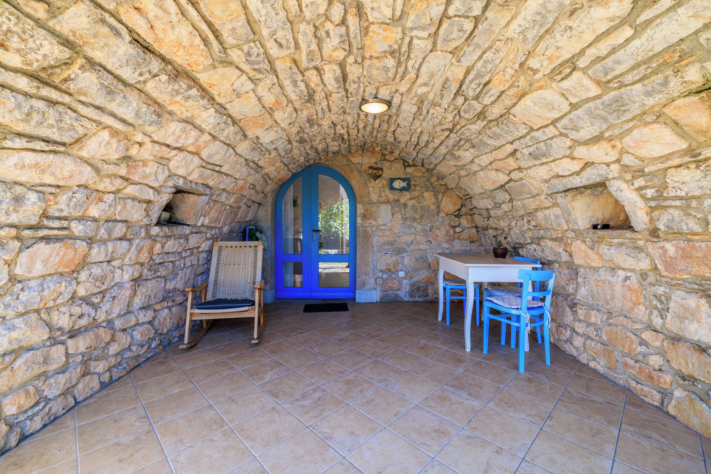 Une pièce aux murs de pierre, dotée d’un sol carrelé, d’une porte bleue et d’une table entourée de chaises.