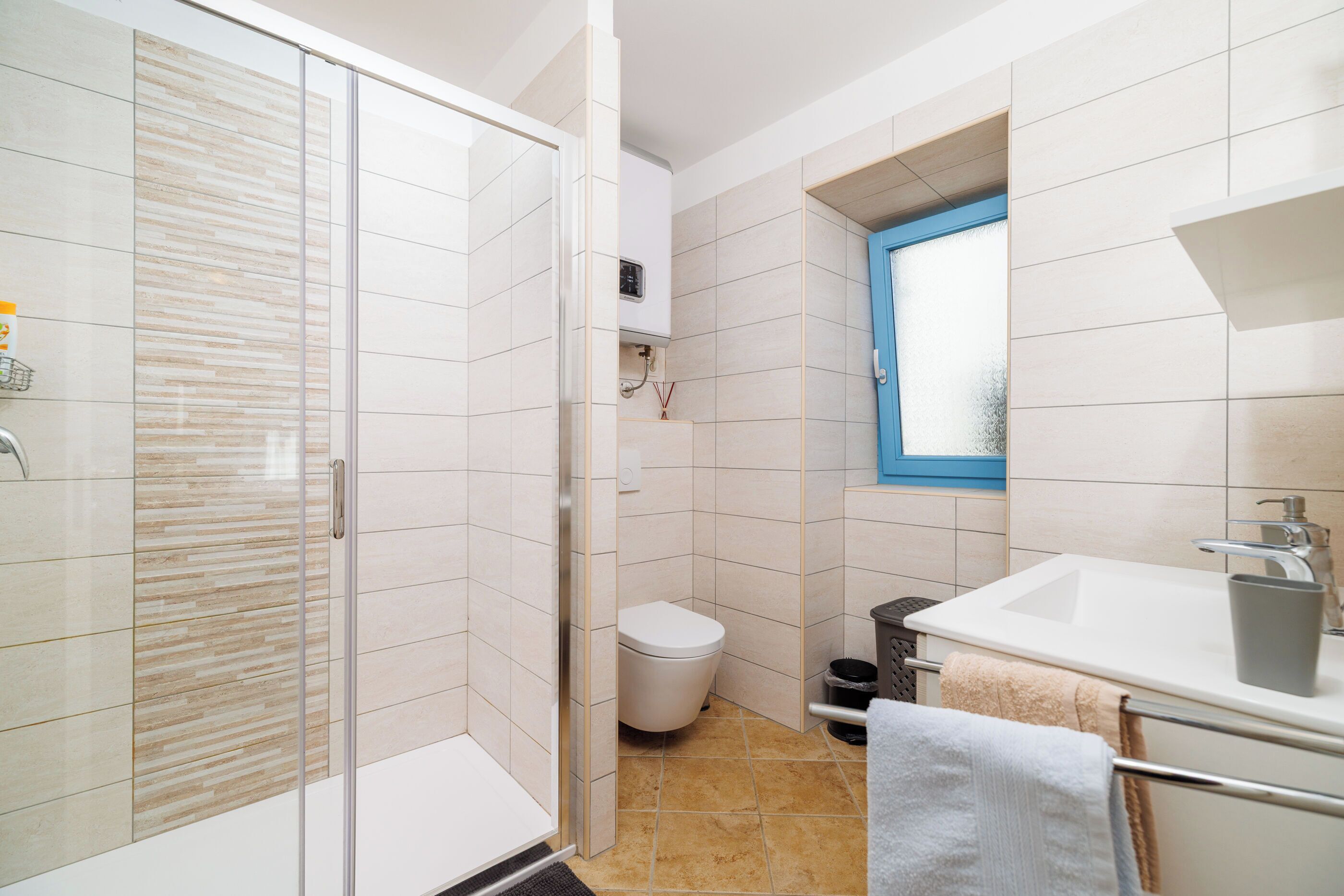 A modern bathroom with a glass shower enclosure, a white toilet, a sink with a towel rack, and a window above the sink.