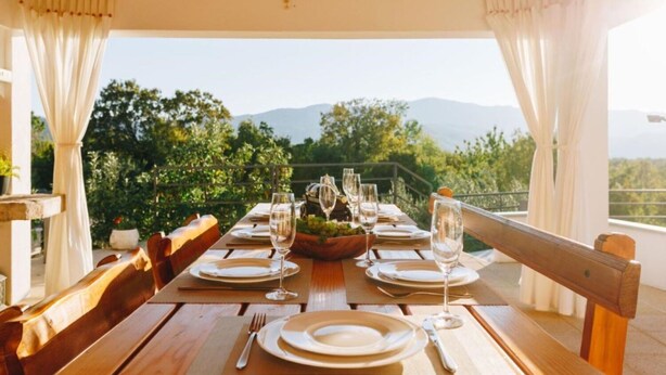 A dining table set for six with white plates, silverware, and wine glasses, overlooking a mountainous landscape. - House Jurica (Omis)