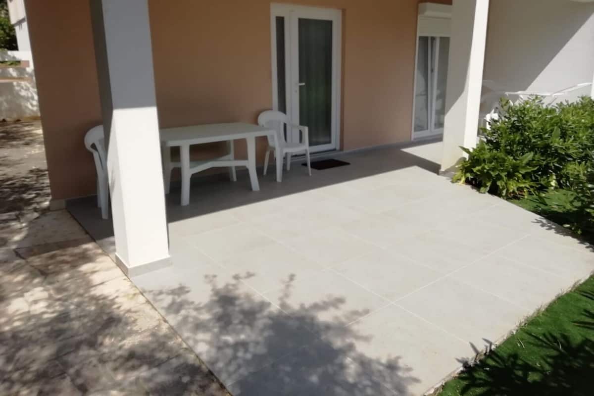 Uma área externa coberta com mesa, cadeiras, uma porta de correr de vidro e piso ladrilhado.