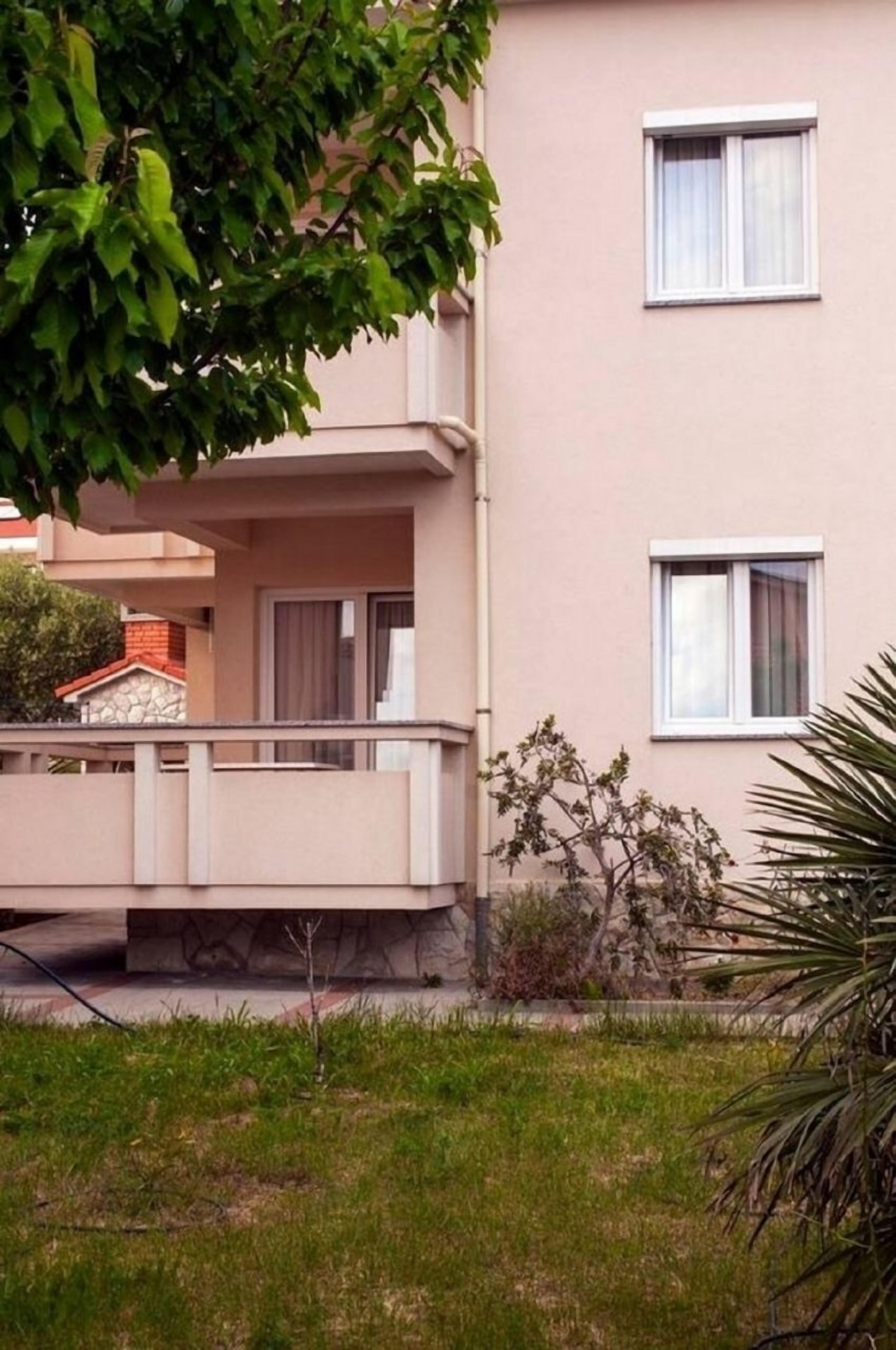 Un edificio de dos pisos con un balcón, un pequeño jardín y un árbol en primer plano.