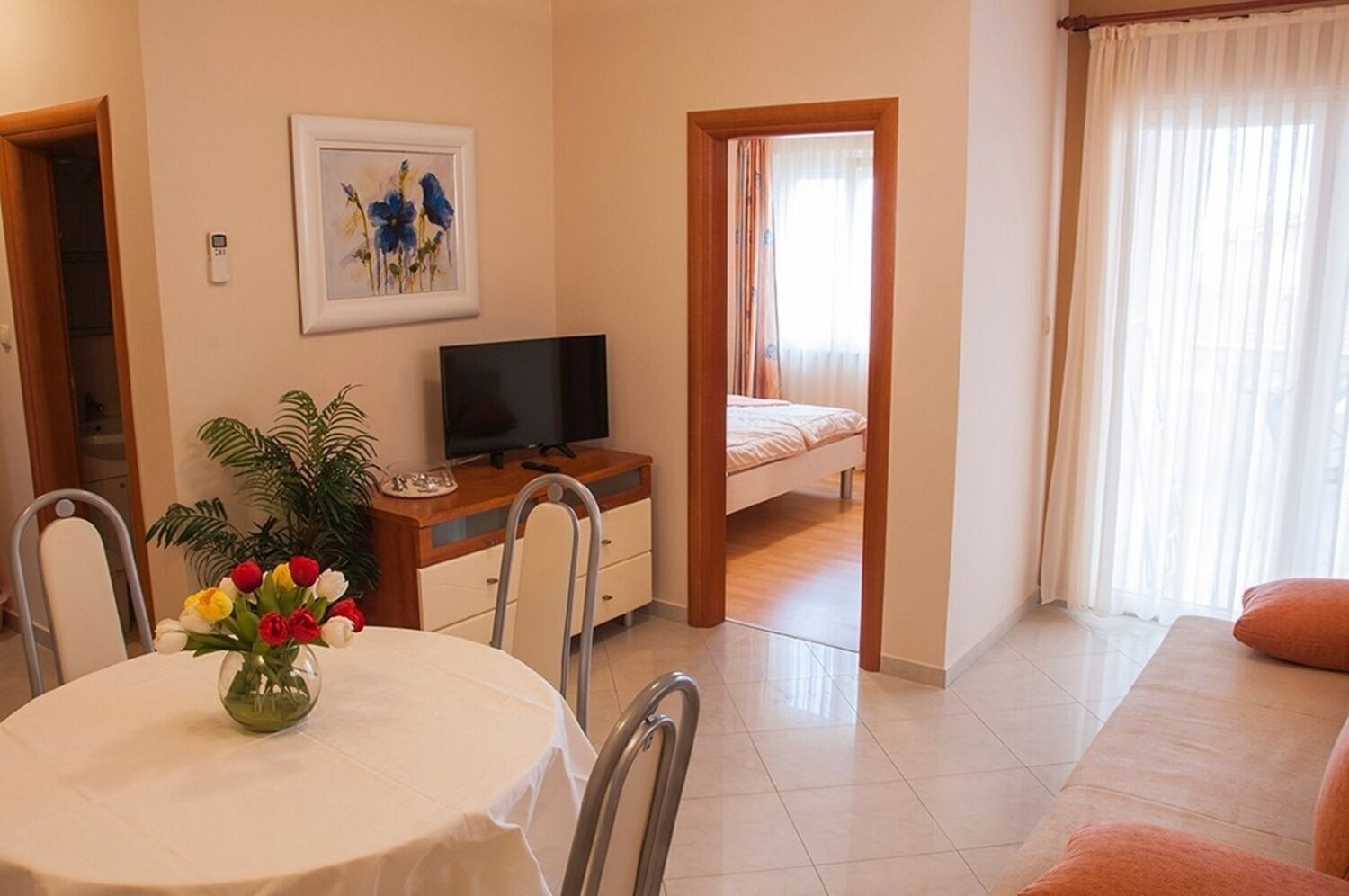 Ein Zimmer mit Esstisch, Stühlen, Fernseher, Bett und einem Fenster mit Vorhängen.