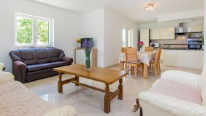 A modern living room with a brown leather sofa, a wooden coffee table, and a white armchair. There is a dining area with a table and chairs, and a kitchen in the background. - House Brist (Drnis)