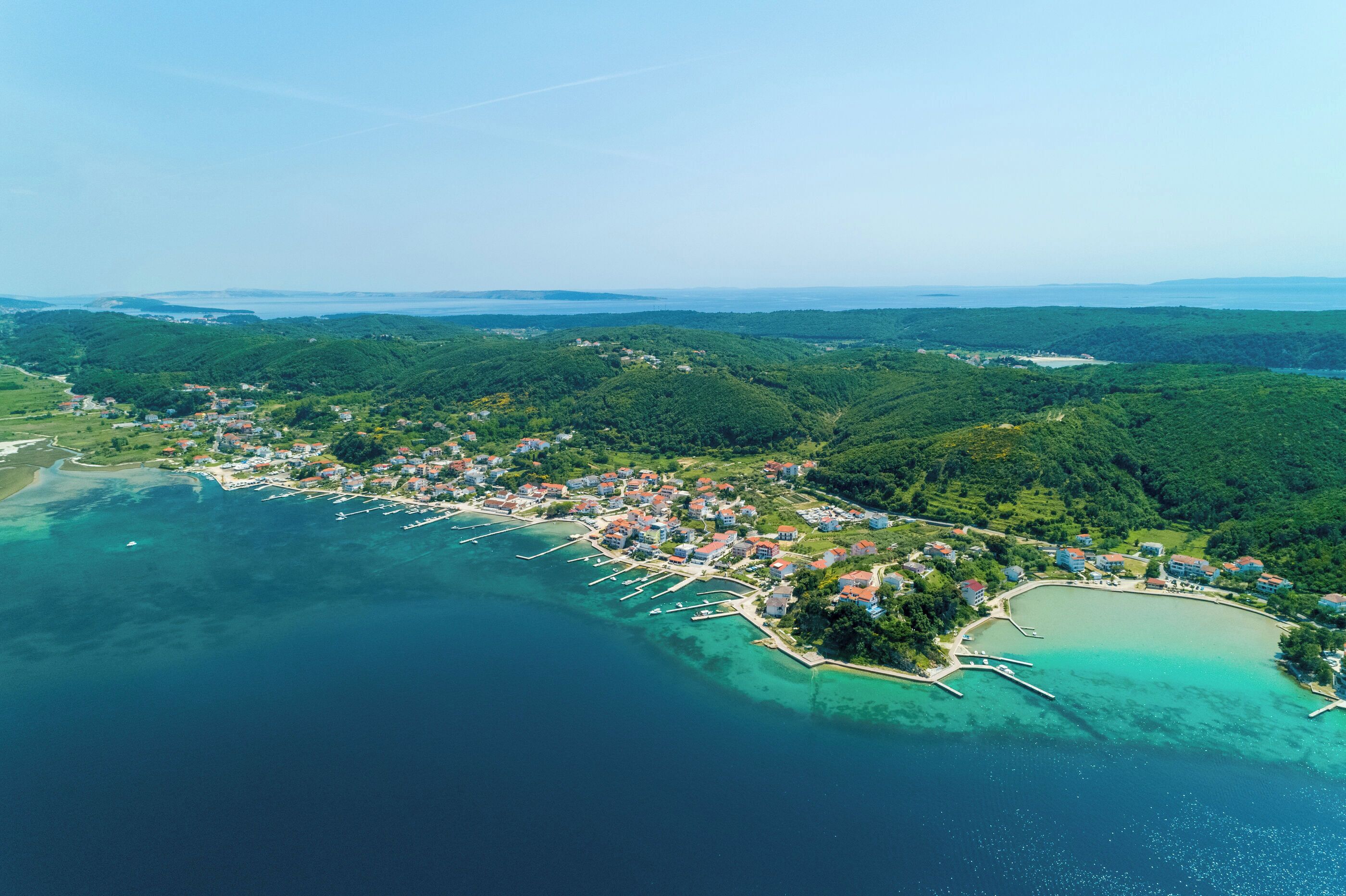 An aerial view of a coastal town with a marina, surrounded by lush green hills and clear turquoise waters.