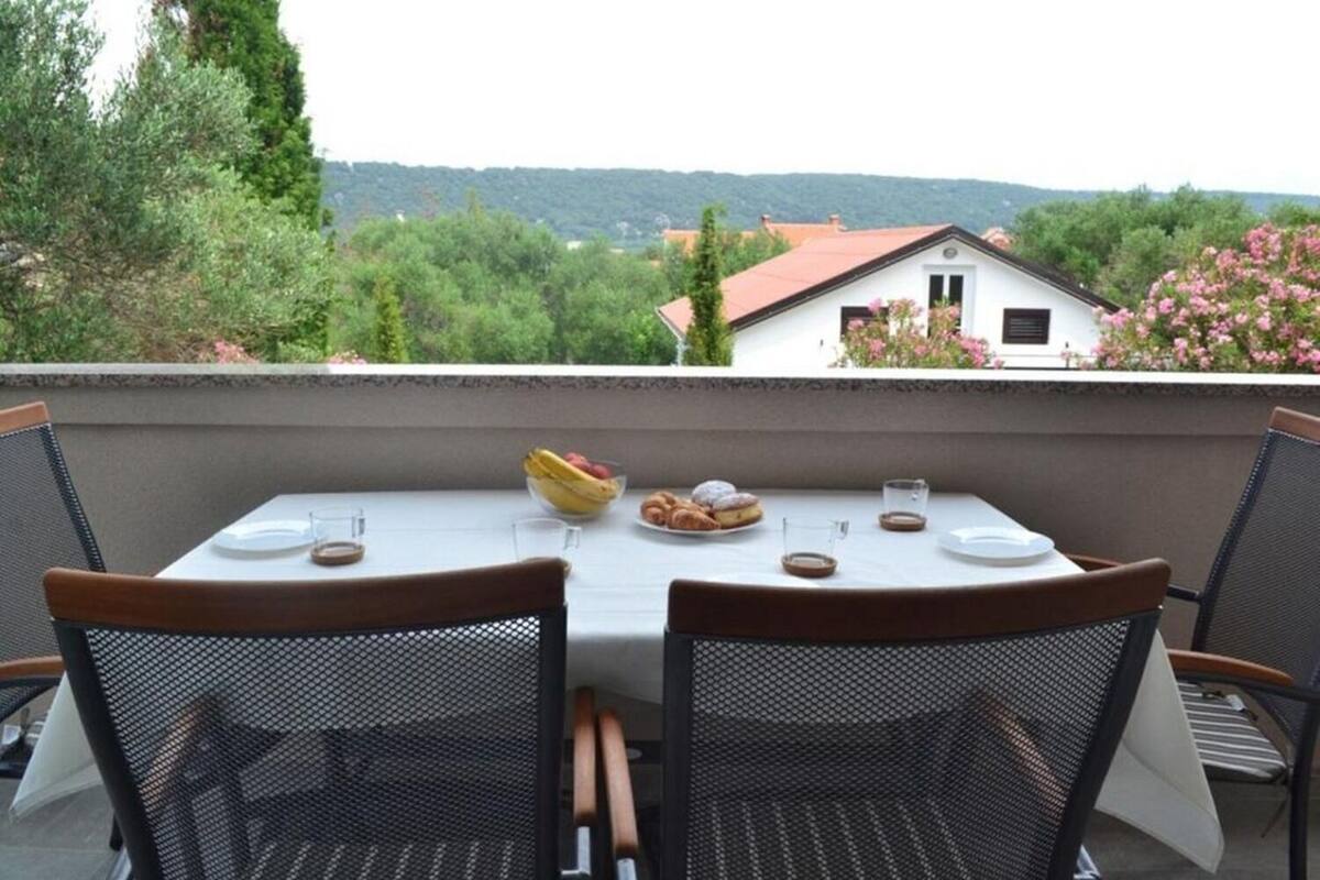 A dining table with chairs on a balcony, overlooking a house and greenery.