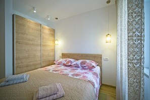 A bedroom with a bed, wooden headboard, two wall-mounted lights, and a patterned curtain. - Apartment Afitojen (Postira)