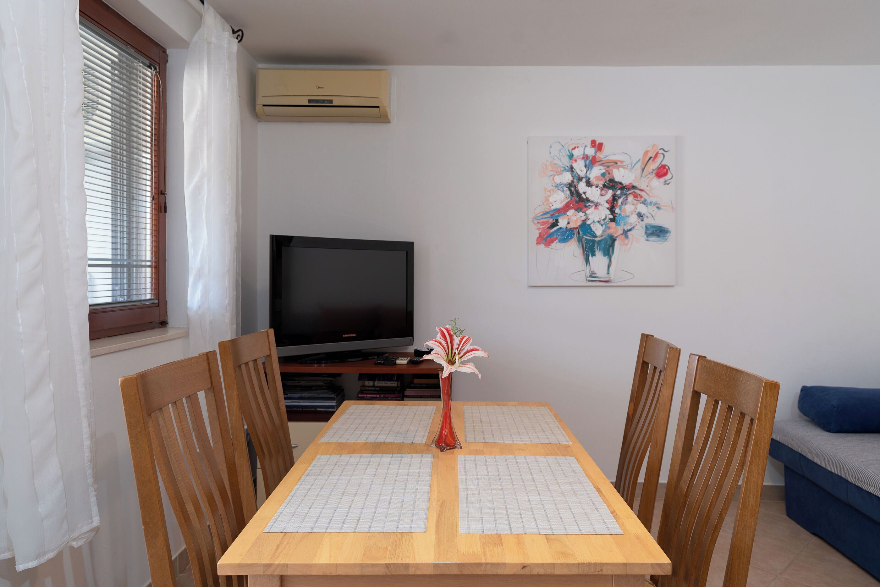 A dining area with a wooden table, four chairs, a vase with flowers, a television, and a painting on the wall.