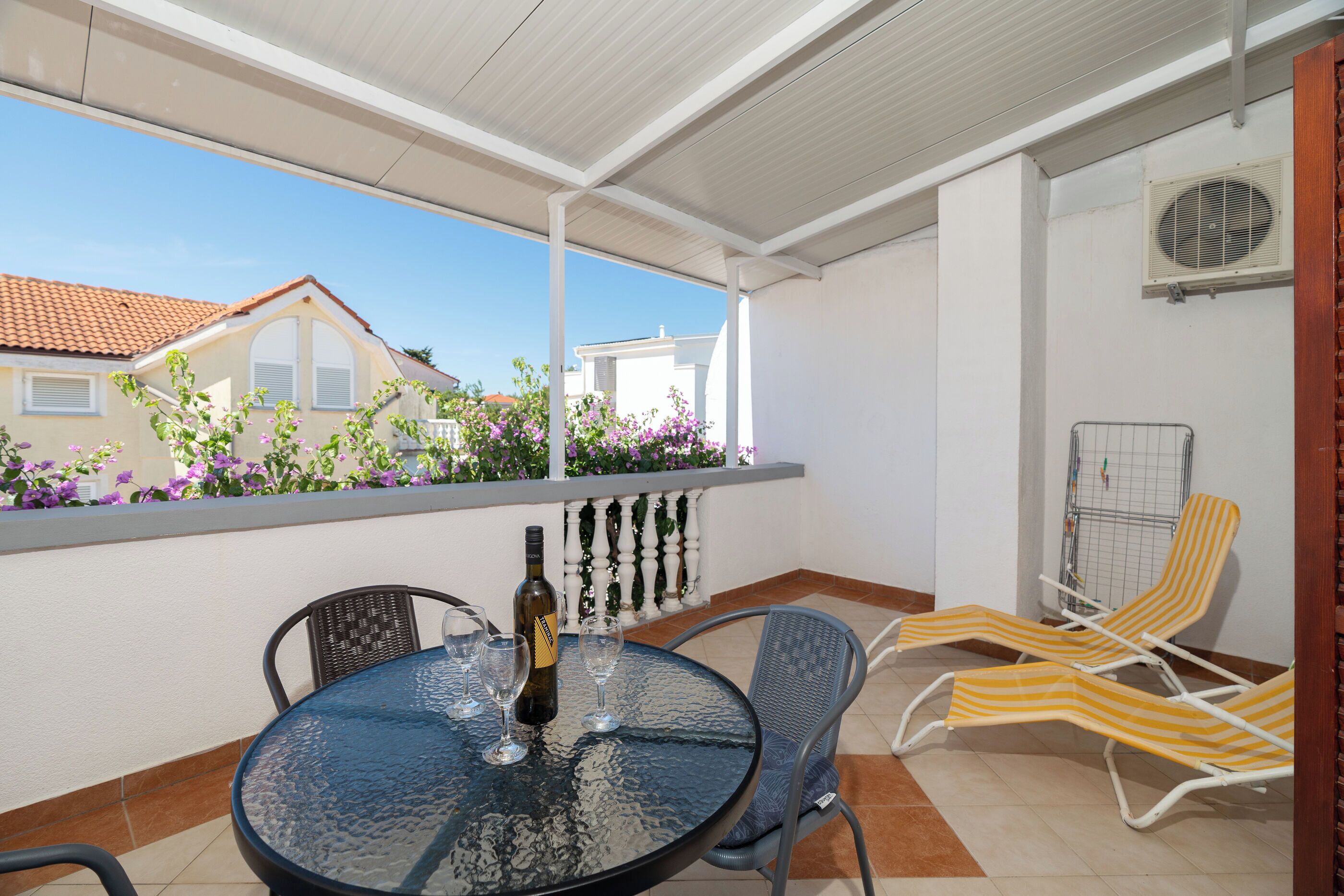 A covered outdoor patio with a round table, chairs, a bottle of wine, and a view of neighboring houses.