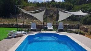 A rectangular swimming pool with clear blue water, surrounded by a stone wall and a paved deck. There are white lounge chairs and a shaded area with a canopy. - House Nane Garden (Supetar)
