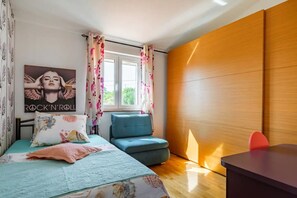 A bedroom with a bed, a blue chair, a wooden wardrobe, and a window with floral curtains. - House Peace (Kastela)