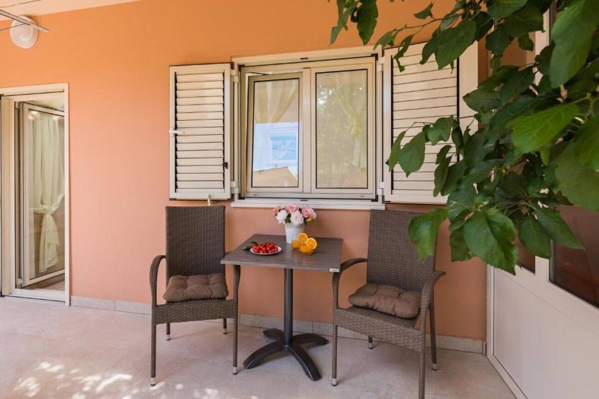 An outdoor seating area with wicker chairs and a table, a window with shutters, and a potted plant.