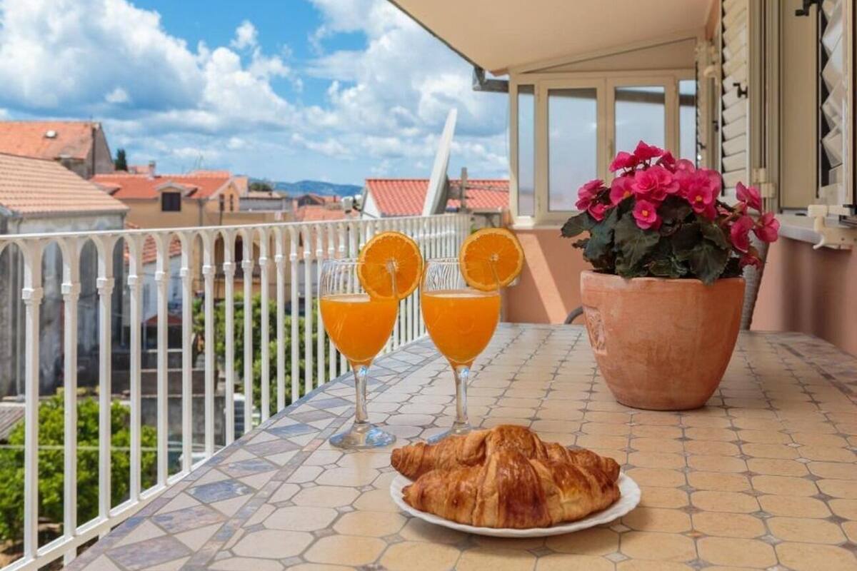 A balcony with a tiled floor, a white railing, a table with a croissant, two glasses of orange juice, and a potted plant with pink flowers.