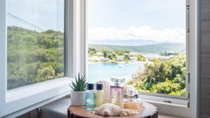 Ein Fenster mit Blick auf eine Küstenlandschaft, ein kleiner Tisch mit Toilettenartikeln und ein Blumentopf.