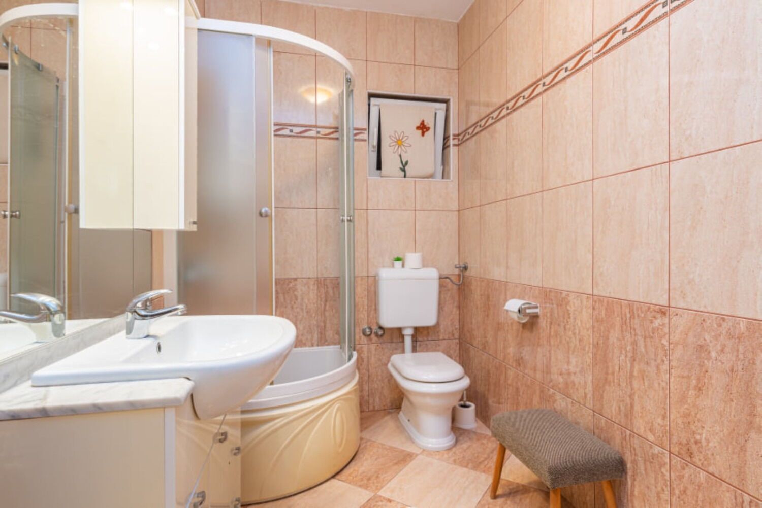 A bathroom with a sink, toilet, shower, and a small stool.