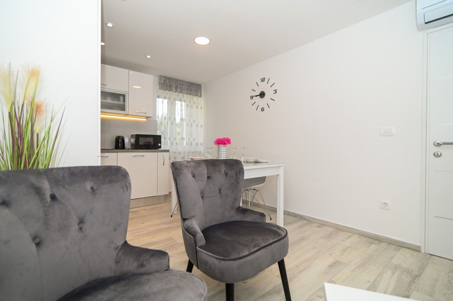 A modern living room with grey upholstered chairs, a dining table, and a kitchen in the background.