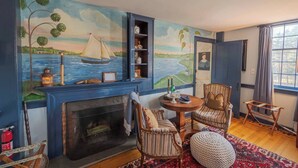 A living room with a fireplace, a round table, two chairs, a rug, and a mural of a sailboat on the wall. - Hospitality as Sweet as Apple Pie! (Stonington)
