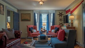 A living room with two red armchairs, a dark sofa, a round ottoman, and a striped rug. There are two windows with blue curtains, a ceiling light, and a wall-mounted ship's wheel decoration. - Hospitality as Sweet as Apple Pie! (Stonington)