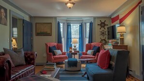 A living room with two red armchairs, a dark sofa, a blue rug, and a round ottoman. There is a window with blue curtains, a wall-mounted ship's wheel, and a lamp on a side table. - Hospitality as sweet as apple pie! (Stonington)