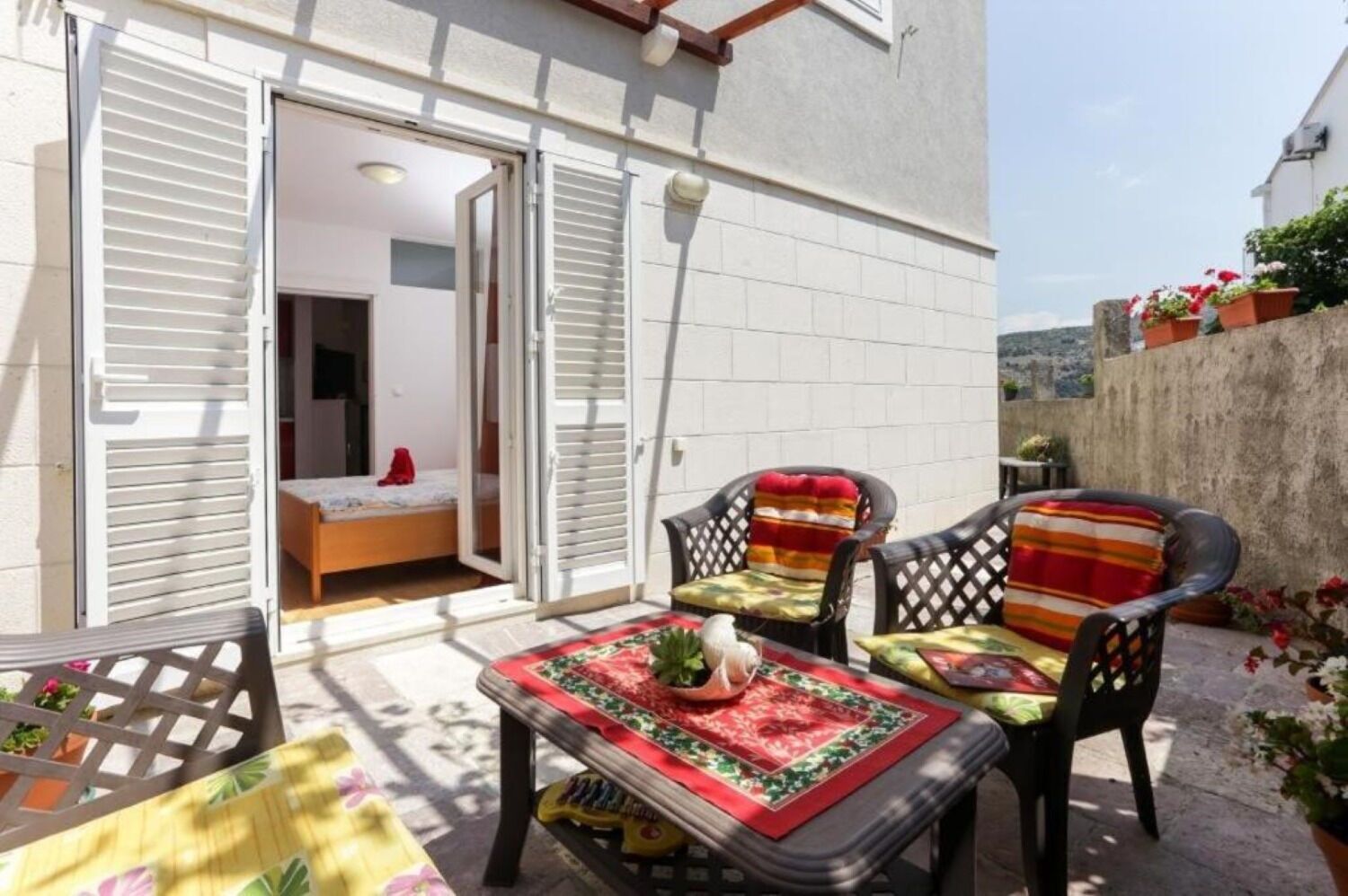 Un patio con una mesa, sillas y plantas en macetas, con una cama al fondo.