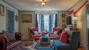 Een gezellige woonkamer met twee rode fauteuils, een blauwe bank, een houten salontafel en een gestreept vloerkleed. Er zijn twee ramen met blauwe gordijnen, een plafondlamp en een decoratieve scheepslamp aan de muur.