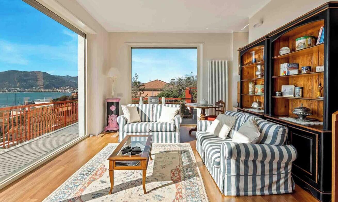Uma sala de estar ampla com uma grande janela que oferece vista para um corpo d'água e colinas, um sofá listrado, uma mesa de centro e uma estante de madeira.