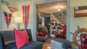 A living room with a blue sofa, a red armchair, a wooden ship wheel, and a flag on the wall. - A Mystic Stay as Sweet as Apple Pie! (Stonington)