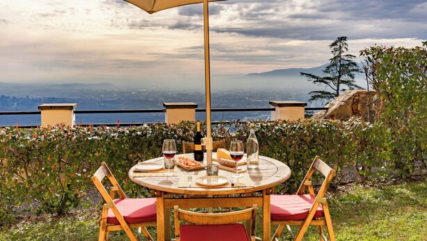 A round table with a beige umbrella, surrounded by wooden chairs with red cushions, set against a scenic backdrop of mountains and a city. - Executive apartment in Borgo di Matraia (Matraia)
