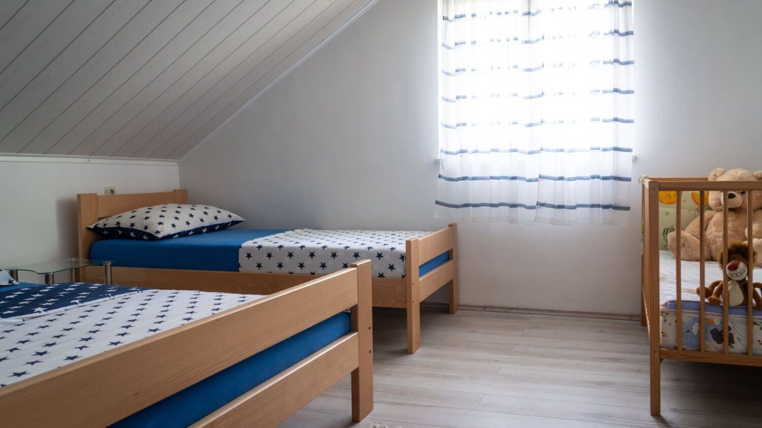 A room with two wooden beds with blue bedding, a crib with toys, and a window with striped curtains.