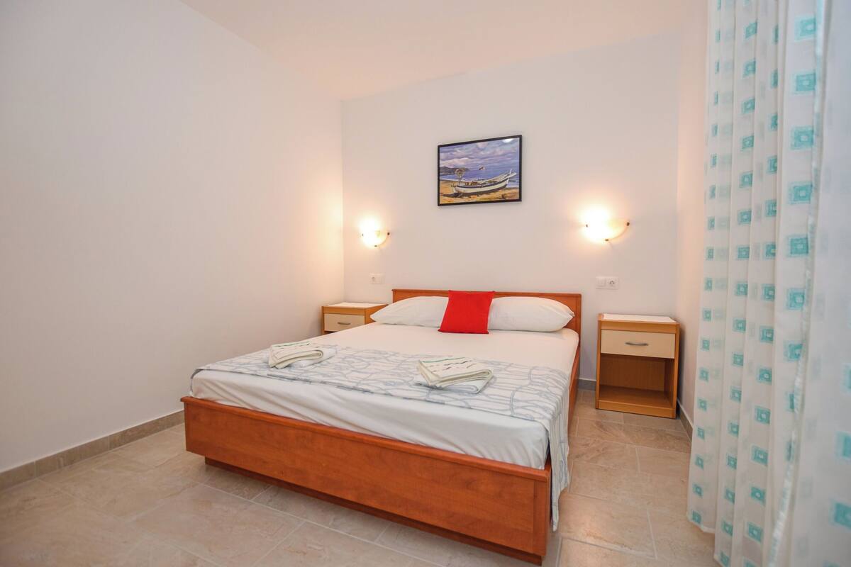 A neatly made bed with a red pillow, two bedside tables, a wall-mounted picture, and a patterned curtain.