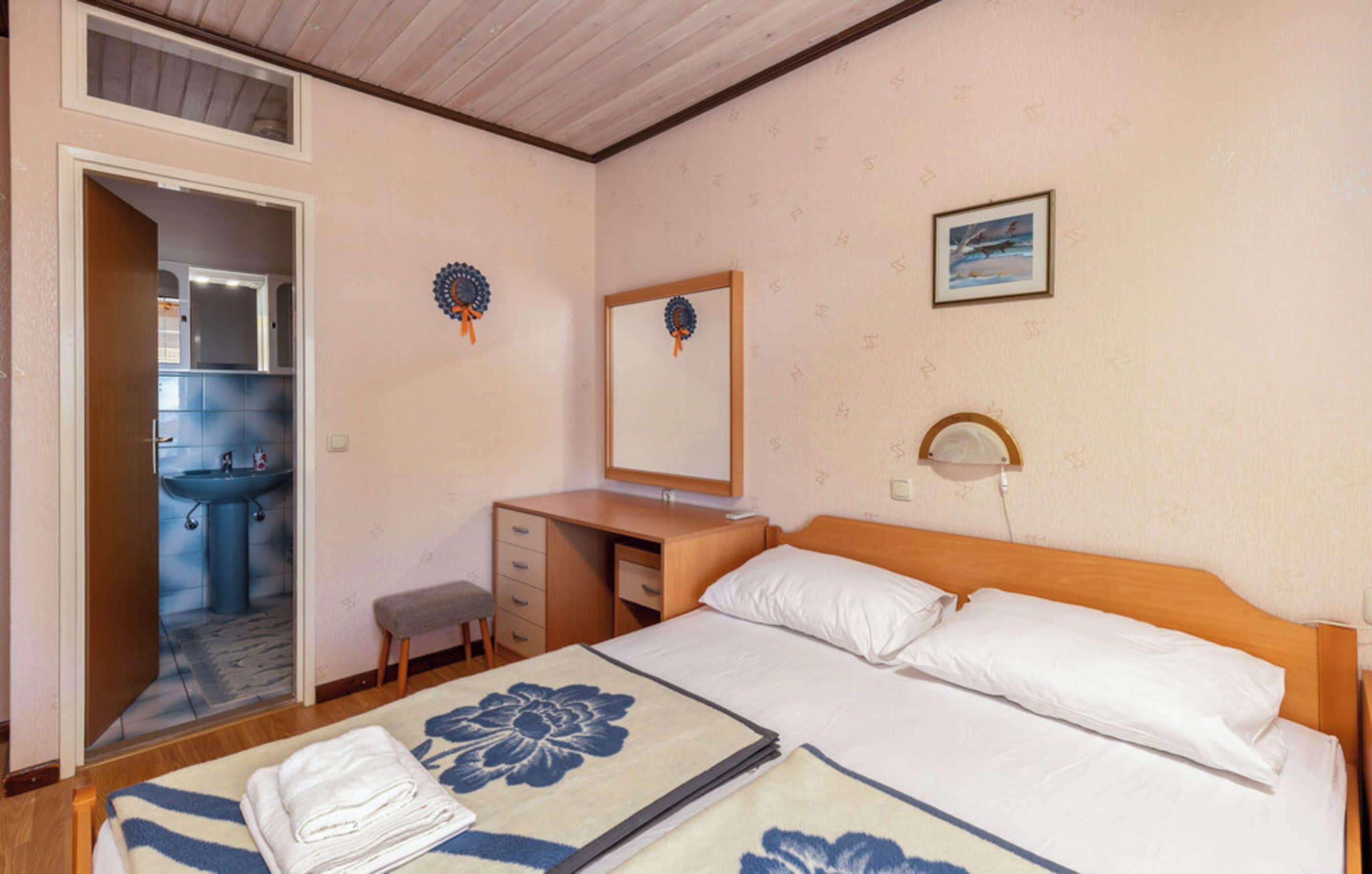 A hotel room with a bed, a desk, a chair, a small stool, a bathroom with a sink, and a mirror.