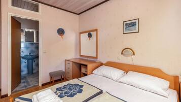A hotel room with a bed, a desk, a chair, a small stool, a bathroom with a sink, and a mirror.