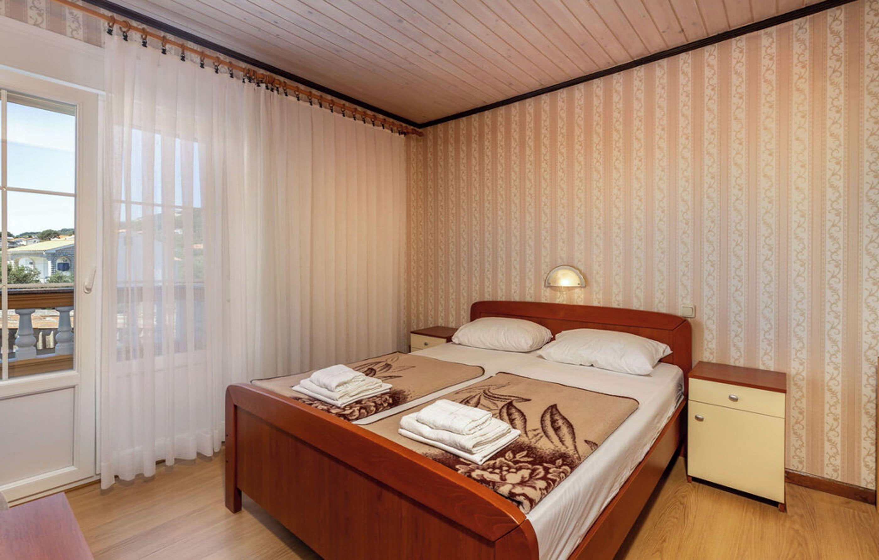 Ein Hotelzimmer mit Bett, Nachttisch, Fenster mit Vorhängen und ornamental gestalteter Tapete.