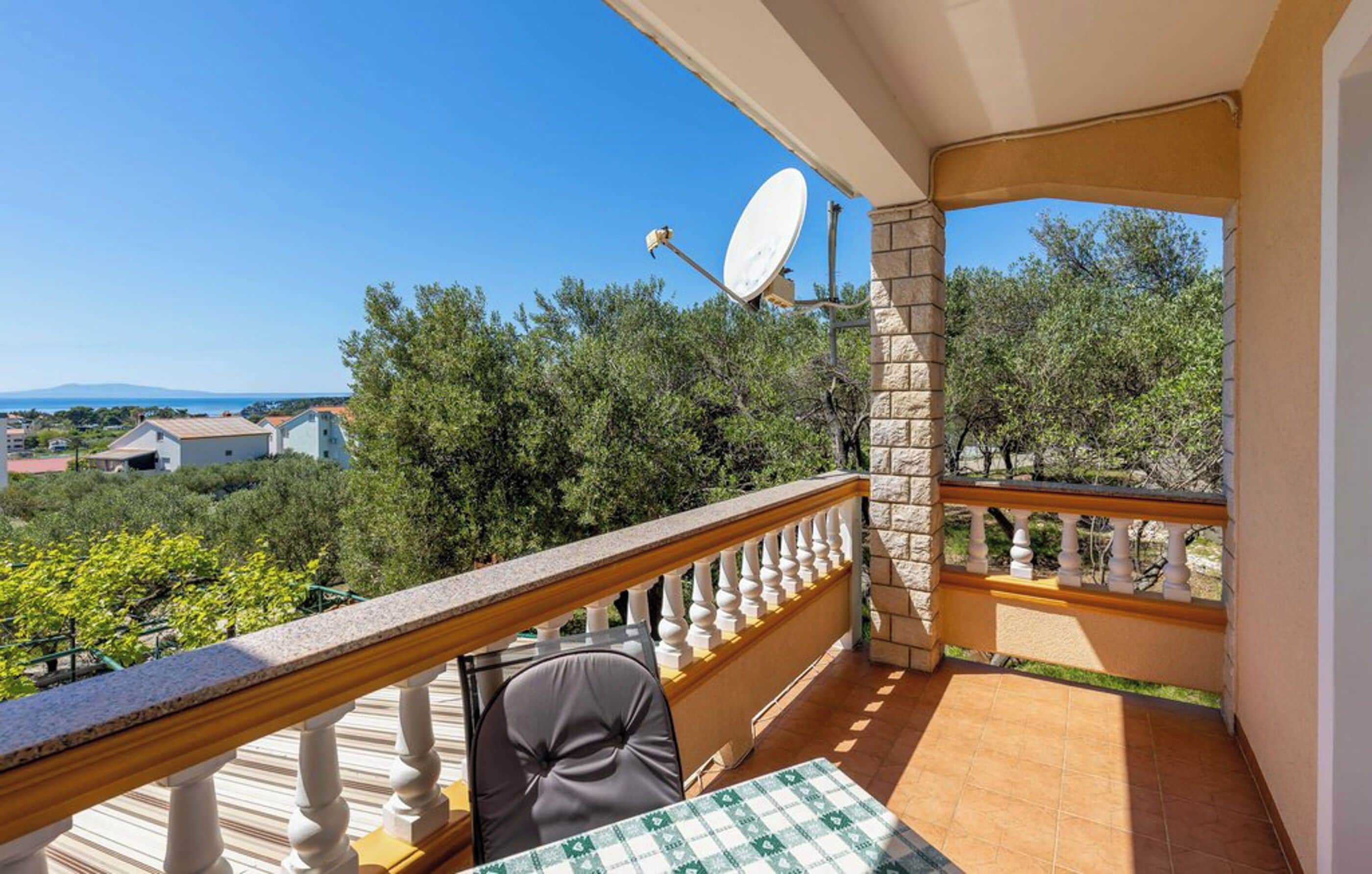 A balcony with a view of trees and buildings, a table with a checkered tablecloth, and a chair.