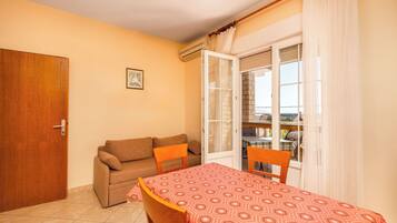 A room with a sofa, a dining table with a patterned tablecloth, a door, and a window with curtains.