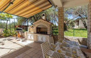 An outdoor patio with a stone barbecue and grill, a table with a placemat, and a pergola overhead. - Apartments Dusan (Rab)