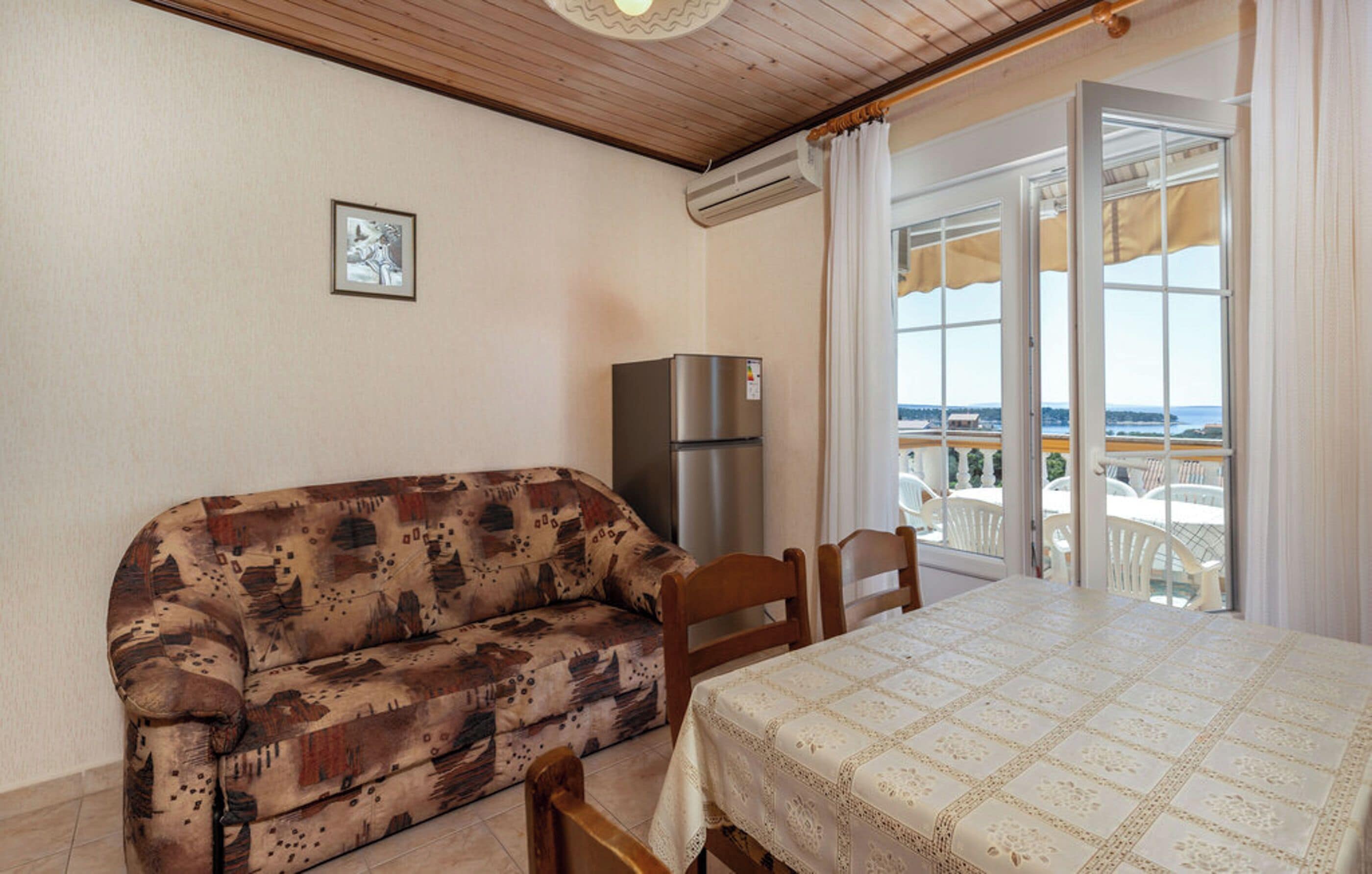 A room with a patterned sofa, a table with a lace tablecloth, a refrigerator, and a view through a glass door.