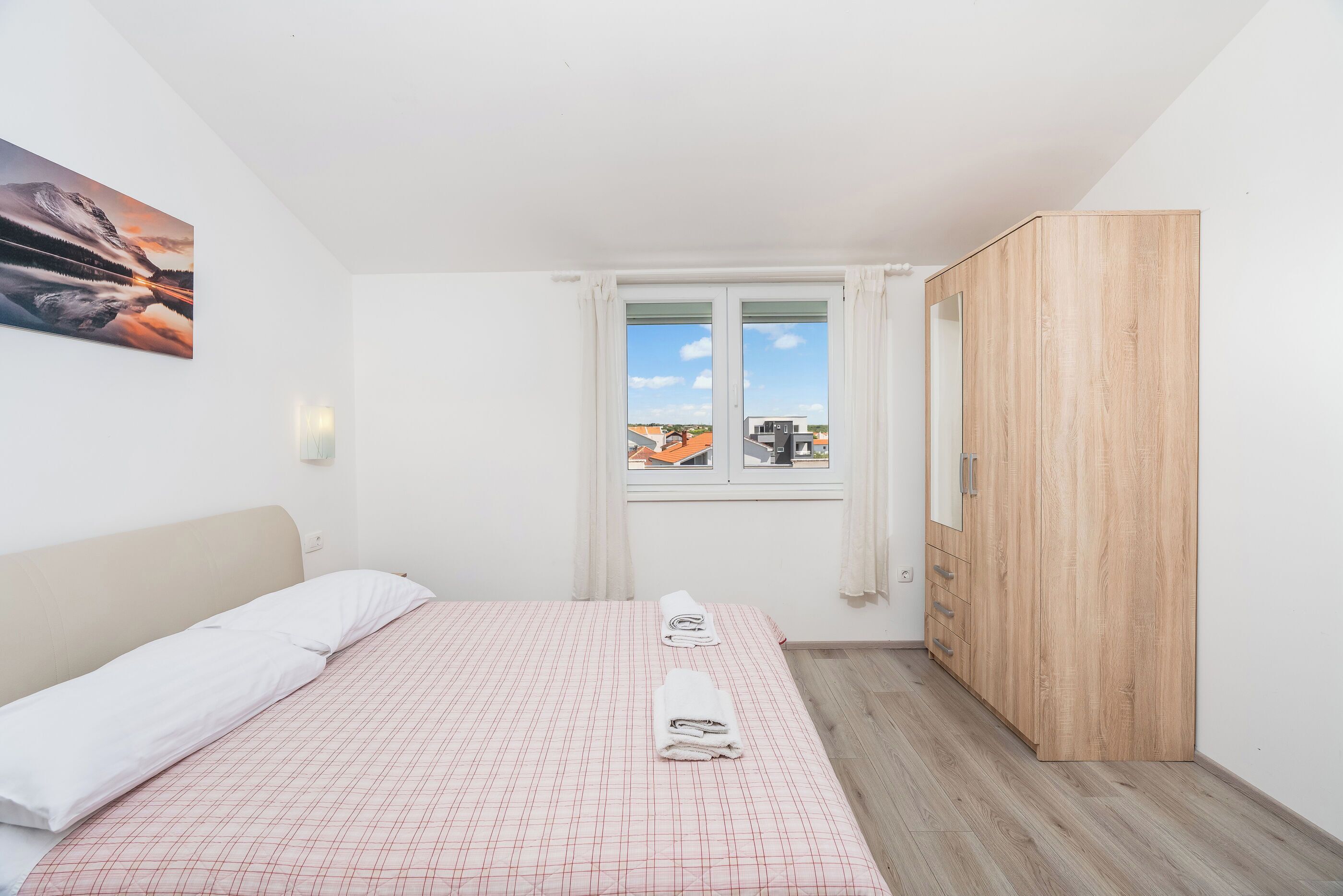 A bedroom with a bed, a window with curtains, a wooden wardrobe, and a painting on the wall.