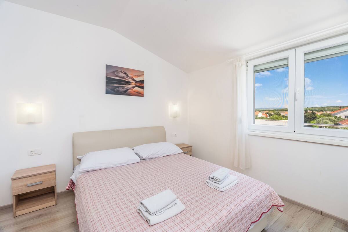 A bedroom with a bed, two bedside tables, a nightstand, a window with a view, and a wall-mounted light fixture.