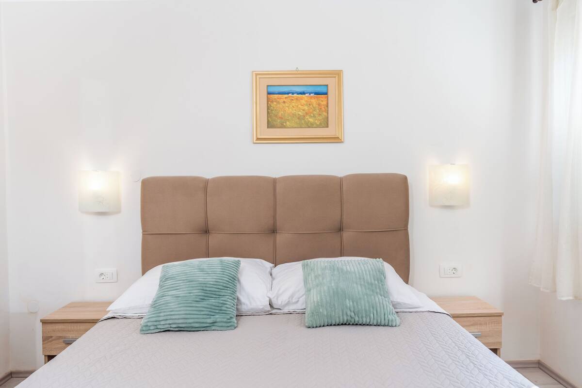 A bedroom with a tufted headboard, two bedside lamps, a framed landscape painting, and two green throw pillows.